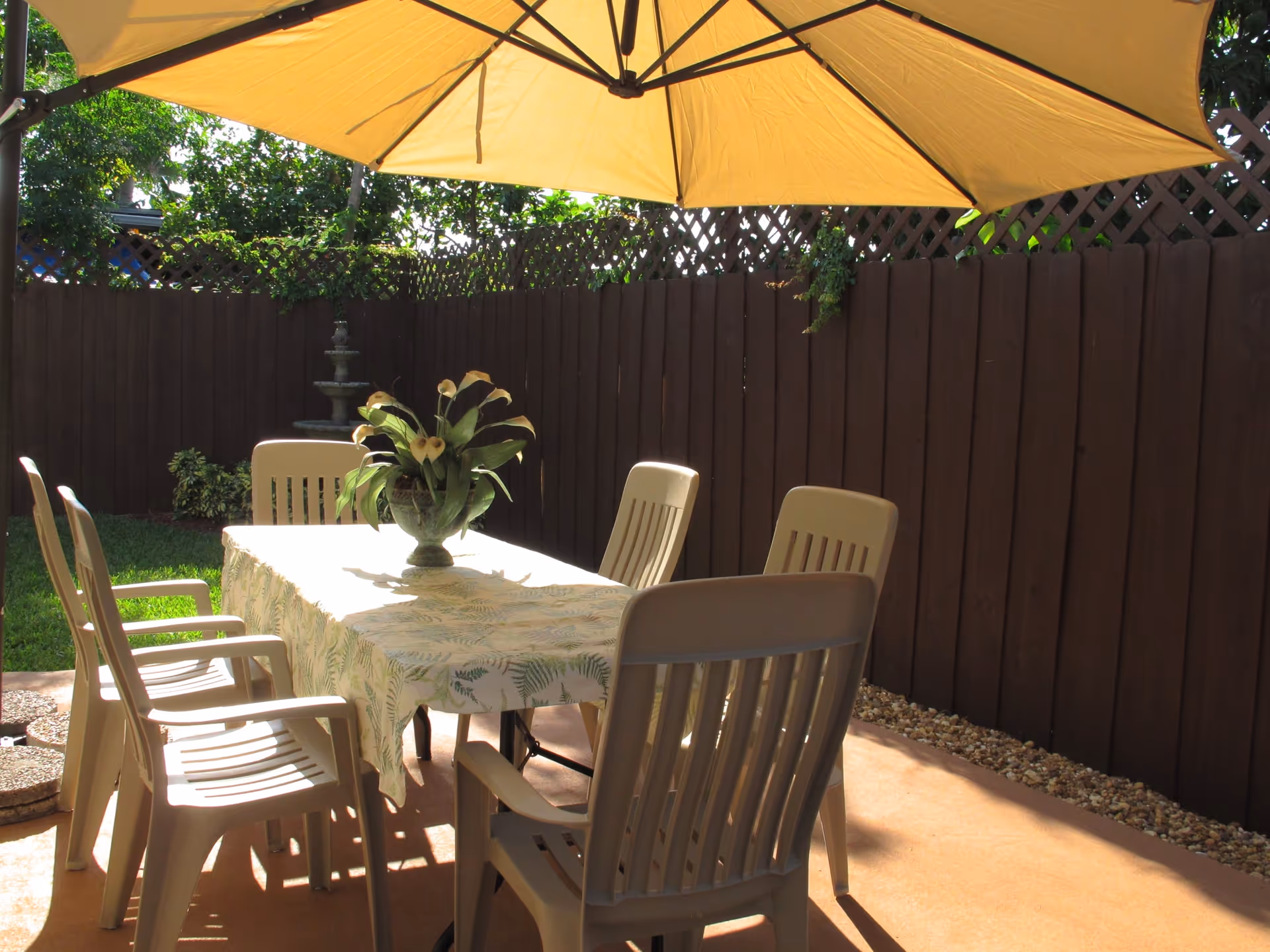 Outdoor patio area with a rectangular table covered by a light-colored tablecloth with a leaf pattern. Six beige plastic chairs surround the table. A large yellow umbrella provides shade. A dark wooden fence encloses the area, with greenery visible above the fence and a small water fountain in the corner.