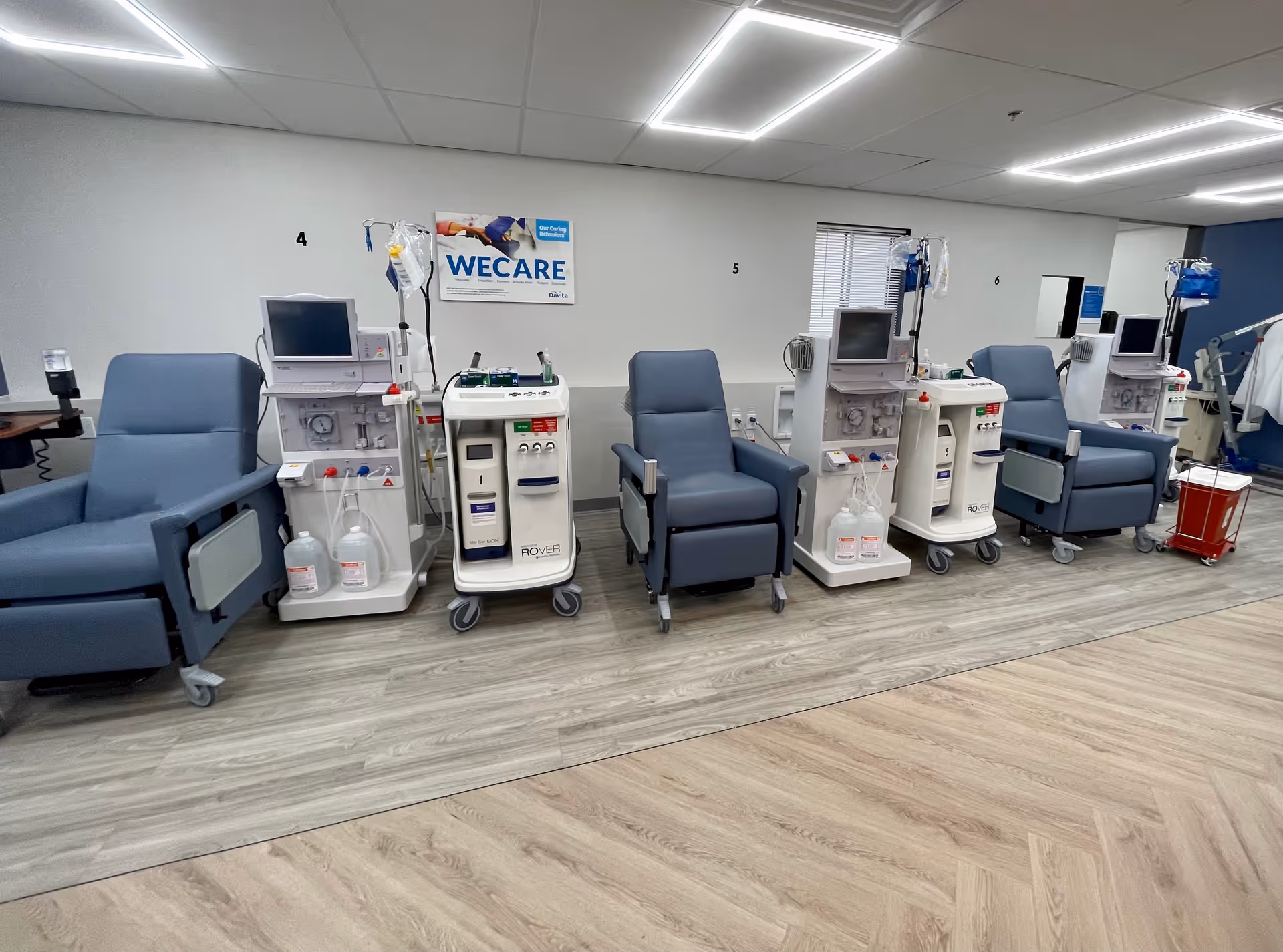 Row of reclining medical chairs and dialysis machines lined up in a treatment room with a 'WECARE' poster on the wall.