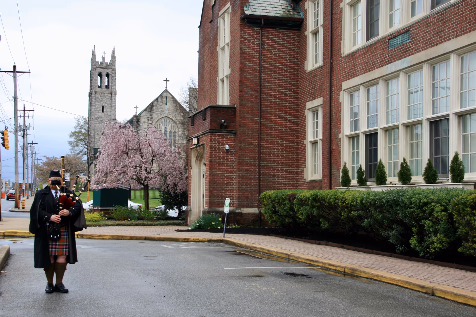 A man dressed in traditional Scottish attire playing bagpipes stands on a street corner near a large brick building with many windows. In the background, there is a stone church with a tall bell tower and a blooming pink tree. The scene appears to be in an urban area with power lines and traffic lights visible.