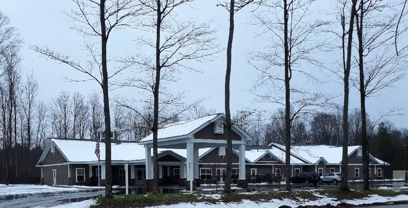 Exterior view of a single-story senior living facility building named August Haus during winter. The building has a covered entrance with white pillars, multiple windows, and a snow-covered roof. There are leafless trees in the foreground and a few parked cars near the building.