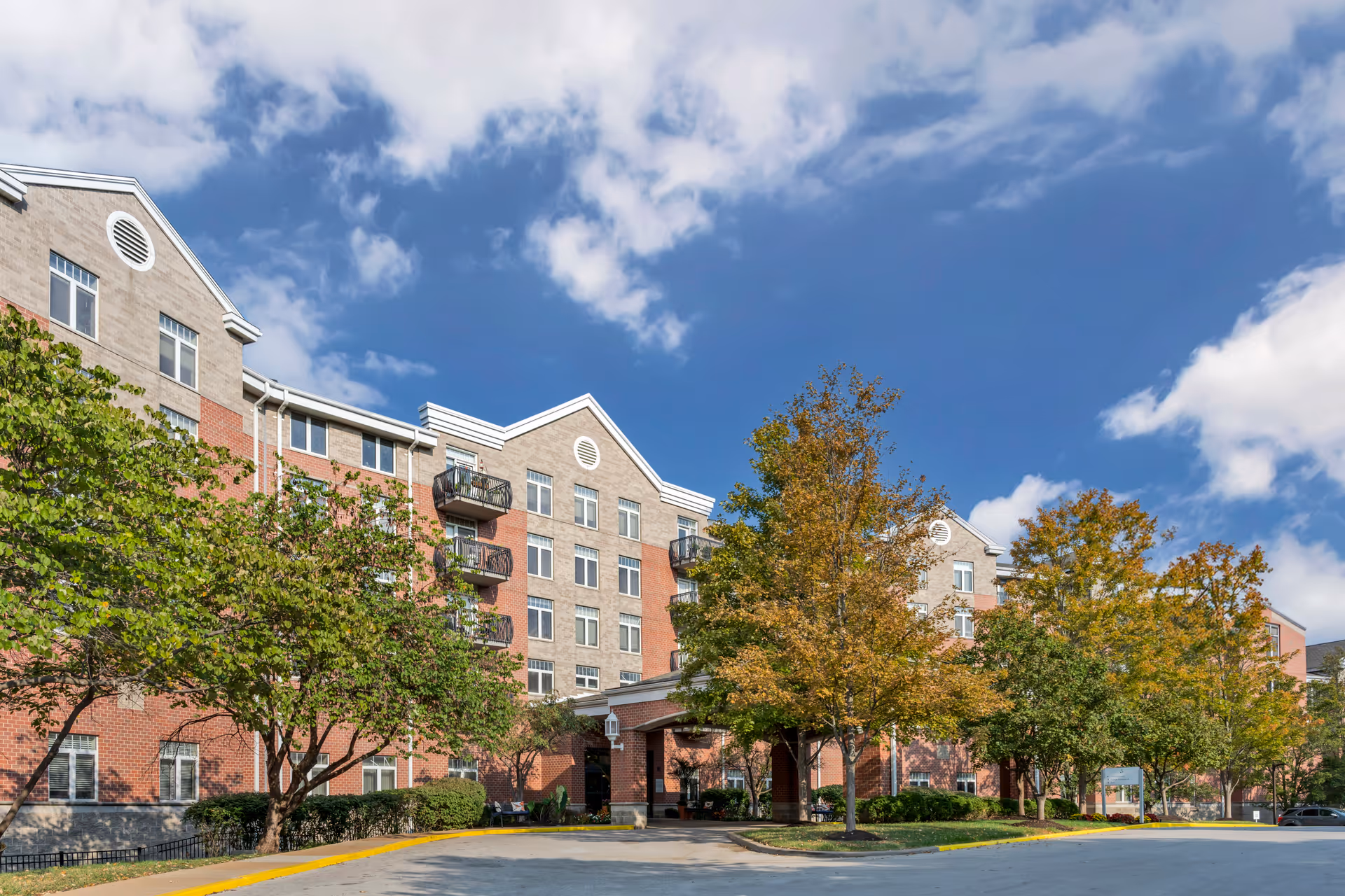 Exterior view of a multi-story senior living facility building with a brick and beige facade, several windows, and small balconies. The entrance is covered with a portico, surrounded by trees with green and autumn-colored leaves under a partly cloudy blue sky.