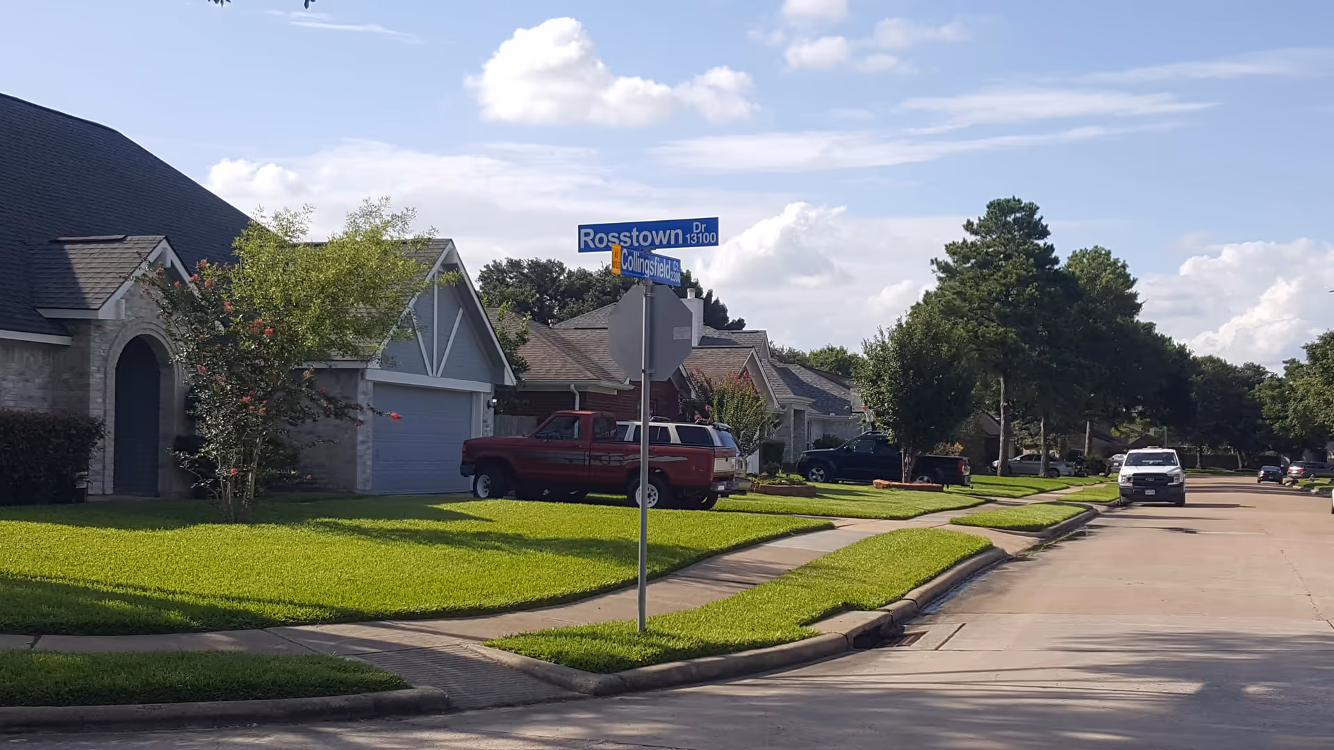 Suburban residential street showing several single-family homes, green lawns, parked vehicles, and a Rosstown Dr street sign under a blue sky.
