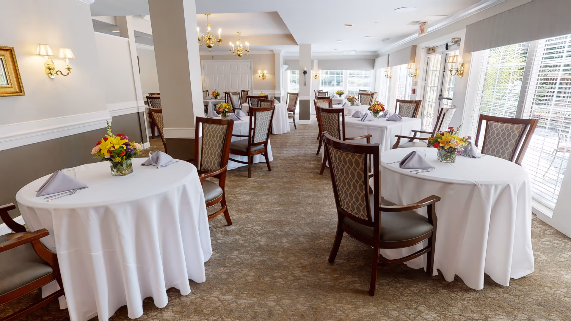 Spacious, well-lit dining room with round tables draped in white tablecloths, floral centerpieces, and upholstered chairs beside large windows.