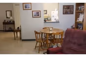 Interior view of a senior living facility showing a small dining area with a round wooden table and four chairs. In the foreground, there is a cushioned armchair. The background includes a kitchen area with a refrigerator and cabinets, and a hallway with a mirror and framed pictures on the walls.