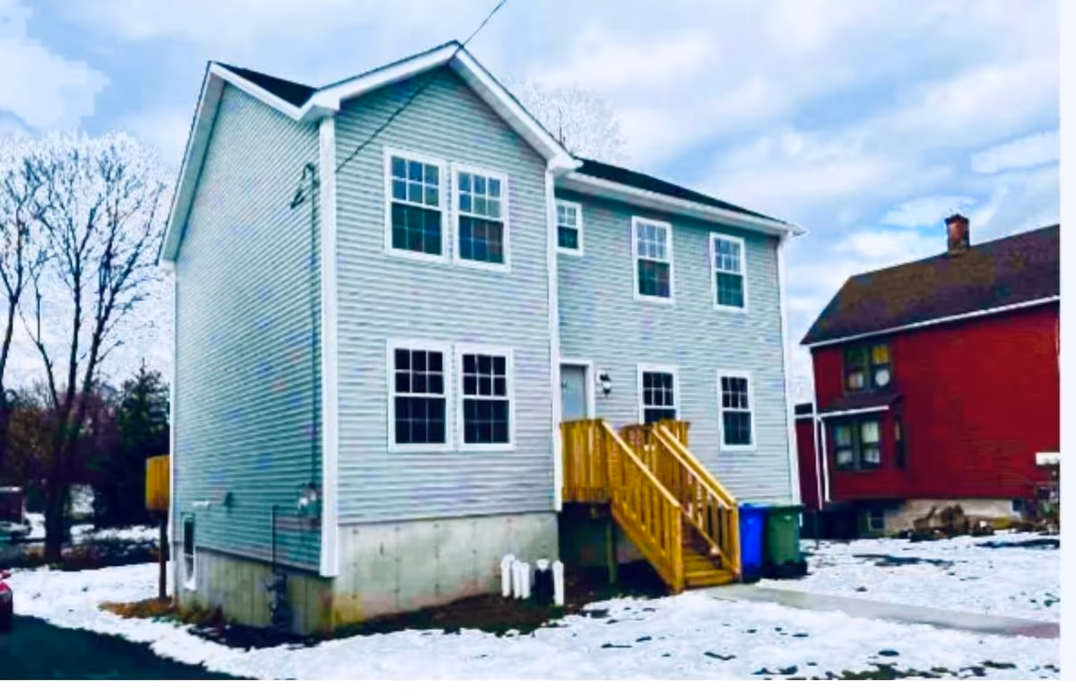 Exterior view of a two-story light gray house with white trim and multiple windows. The house has a small wooden staircase leading to a white door. Snow covers the ground around the house, and there is another red house visible in the background under a cloudy sky.