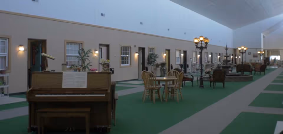Large indoor common area in a senior living facility with a piano, tables and chairs, lampposts, and door-front apartment units along a wide carpeted corridor.