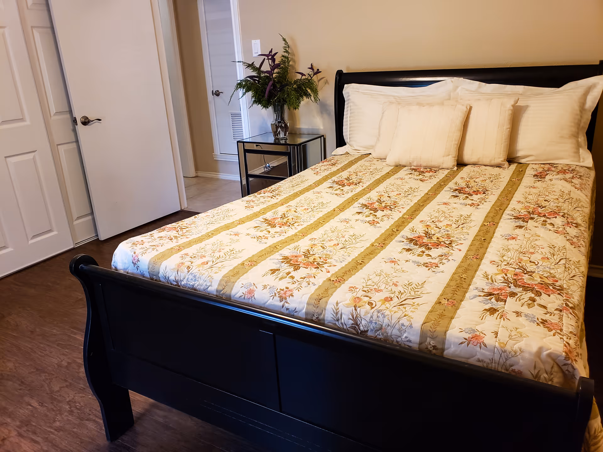 Bedroom with a black wooden bed dressed in a floral bedspread and pillows, a mirrored nightstand with flowers, and an open closet door.
