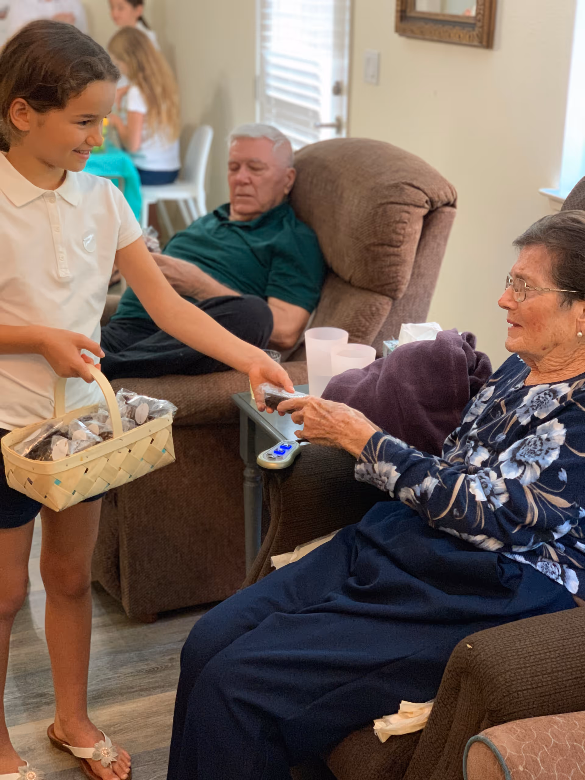 A young girl in a white shirt and shorts is handing a treat from a basket to an elderly woman sitting in a recliner chair. Another elderly man is sitting in a recliner chair in the background, and two other people are seated at a table further back. The setting appears to be a cozy living room with natural light coming through the window.