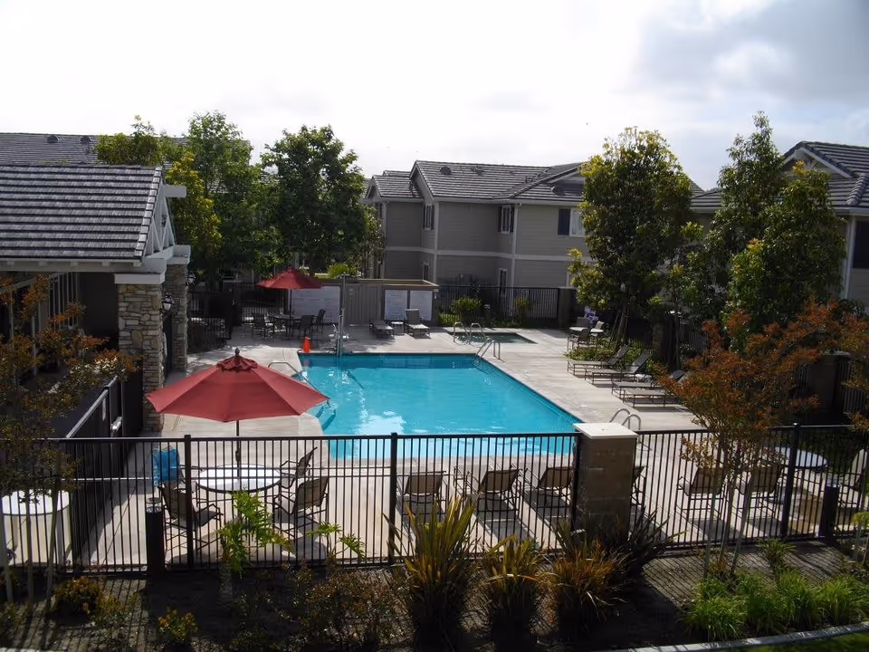Outdoor swimming pool area at a senior living facility with lounge chairs, tables with red umbrellas, surrounded by trees and multi-story residential buildings.