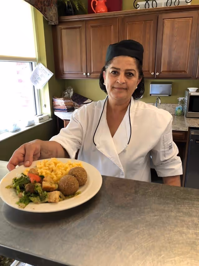 A chef wearing a white uniform and black hat stands behind a counter in a kitchen, holding a plate of food that includes macaroni and cheese, two falafel balls, and a salad with croutons and tomato.