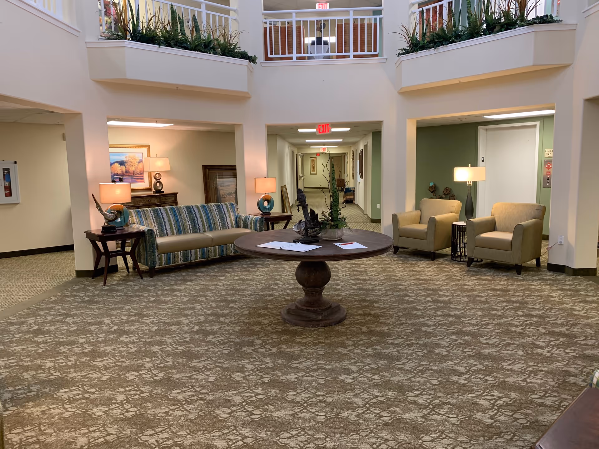 A spacious and well-lit common area in a senior living facility with a patterned carpet, a round wooden table with decorative items and papers, a striped sofa with two side tables and lamps, two beige armchairs with a small table and lamp between them, and a hallway leading to other rooms. There are plants on the upper ledge and exit signs visible in the hallway.