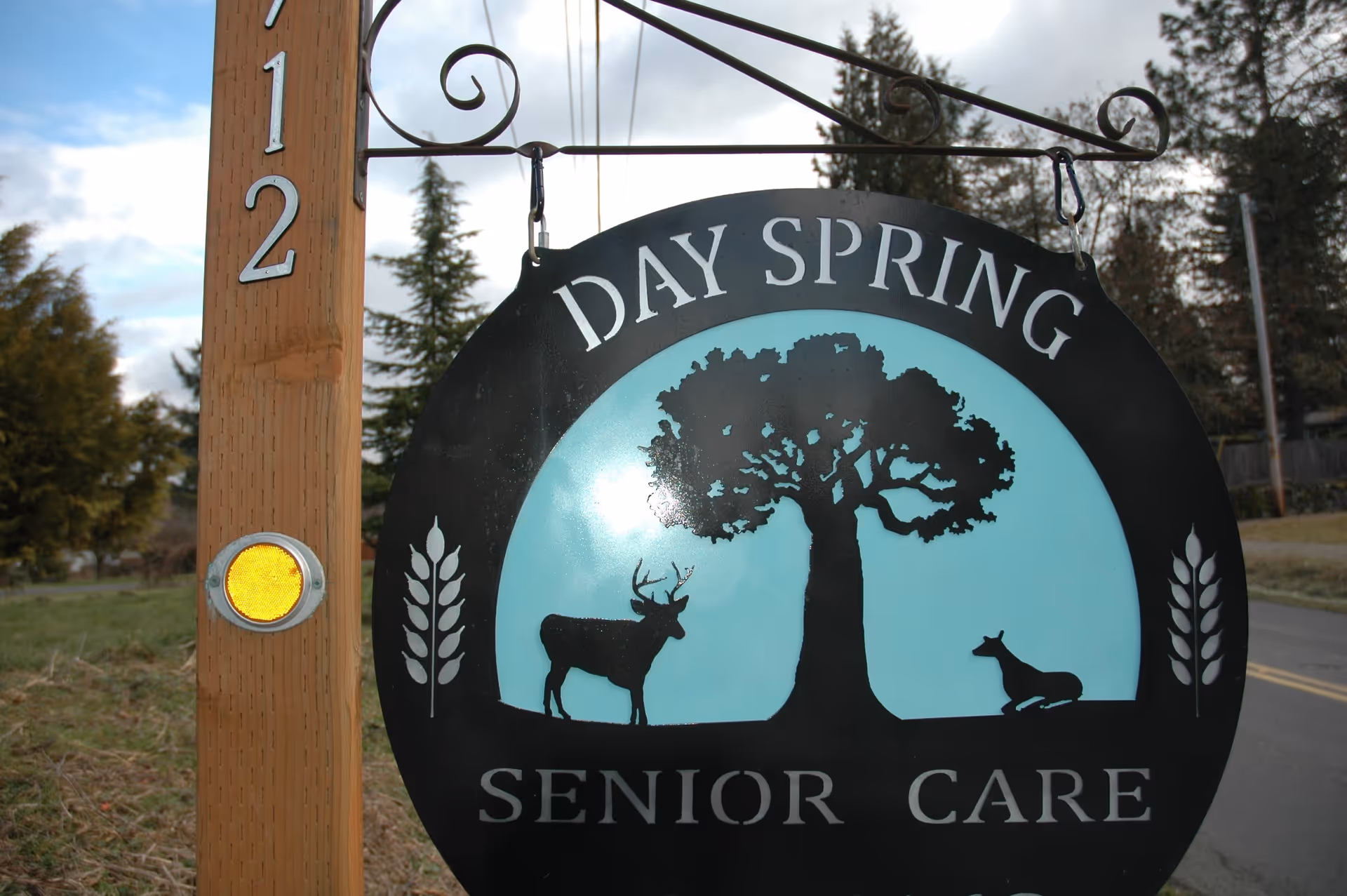 A black and blue sign hanging from a wooden post with the text 'DAY SPRING SENIOR CARE' and silhouettes of a tree, a standing deer, and a resting deer. The post has the number 12 and a yellow reflector attached. Trees and a road are visible in the background.