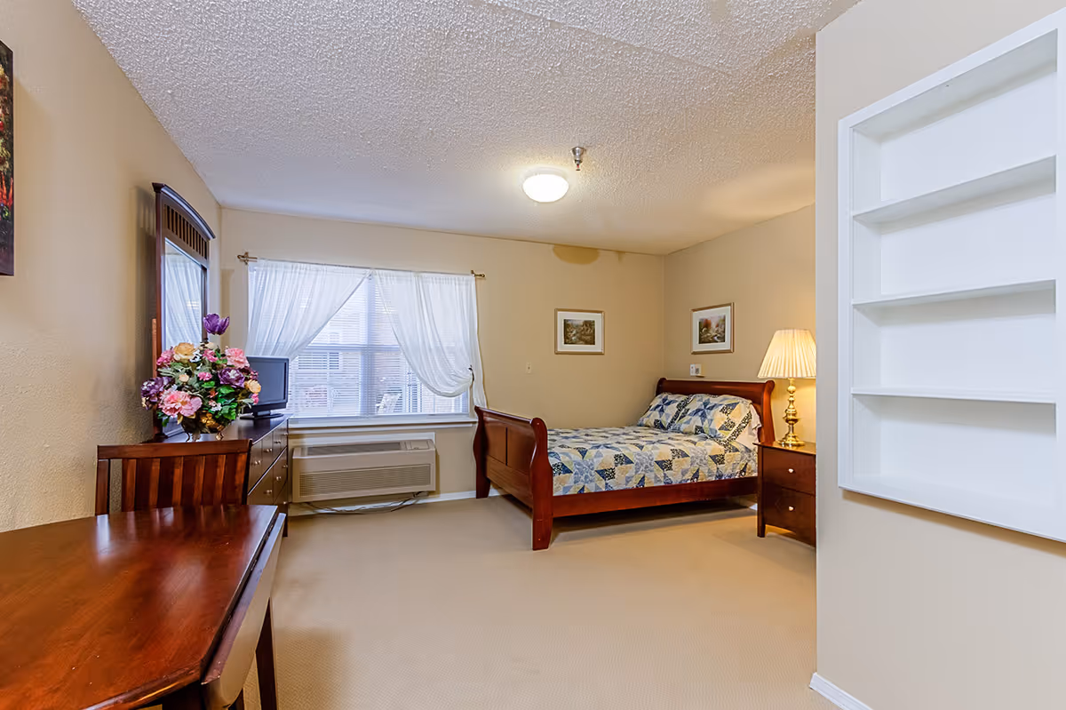 Furnished senior apartment bedroom with a wooden sleigh bed, nightstand and lamp, dresser with flowers, desk, and a curtained window.