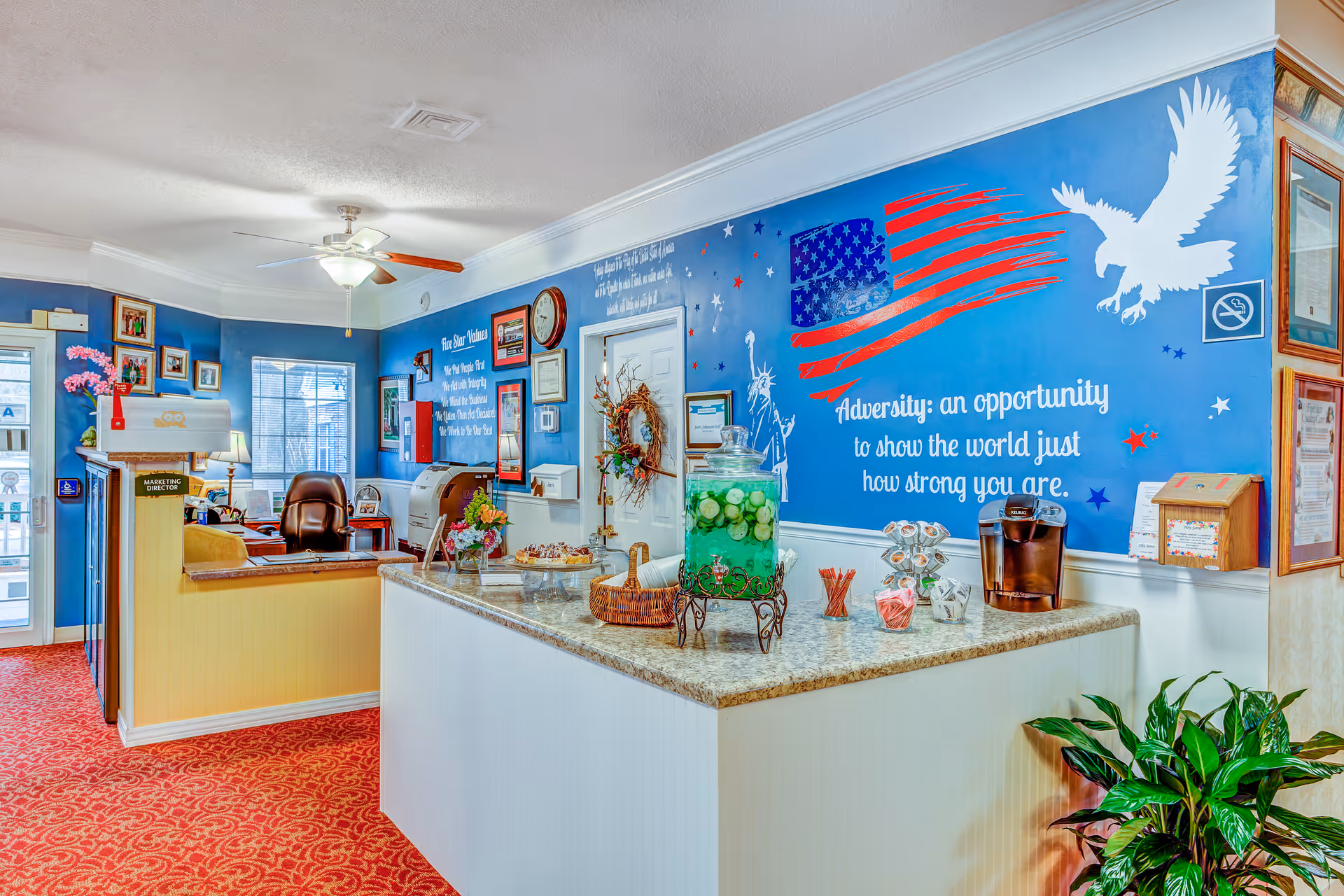Reception area of The Legacy of Orangeburg facility with a front desk, a blue wall featuring an American flag mural, an eagle, and an inspirational quote. The desk has a large glass container with cucumber water, a coffee machine, and various small items. The room has red carpet, framed pictures on the walls, and a ceiling fan with lights.