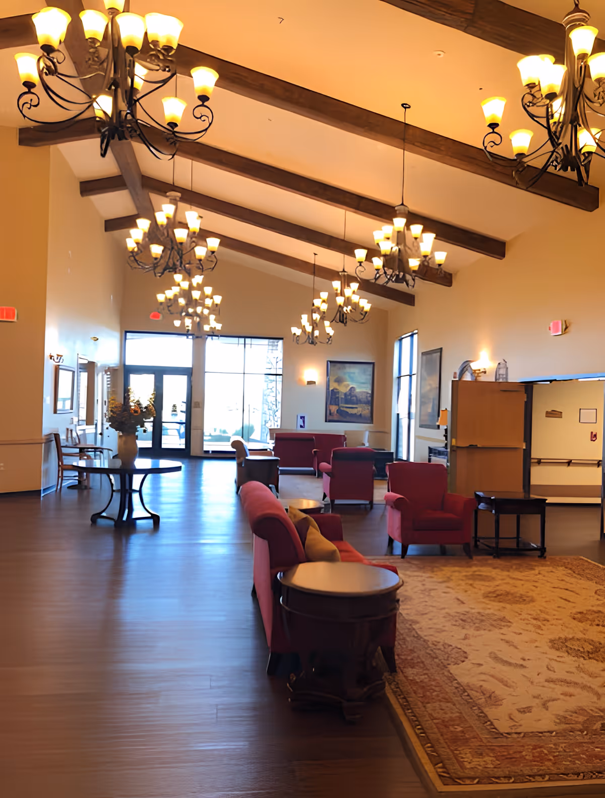 A spacious and well-lit living room area with high vaulted ceilings featuring exposed wooden beams and multiple ornate chandeliers. The room is furnished with red upholstered sofas and armchairs arranged around wooden side tables and a large patterned area rug. Large windows and glass doors allow natural light to enter the space. There are framed paintings on the walls and a table with a vase of flowers near the entrance.