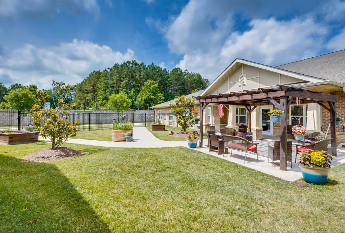Outdoor patio area at Chatham Ridge Assisted Living with a wooden pergola, outdoor seating including chairs and benches, potted plants, a grassy lawn, and a paved walkway. The building exterior is visible with windows and a brick facade, surrounded by trees and a partly cloudy sky.