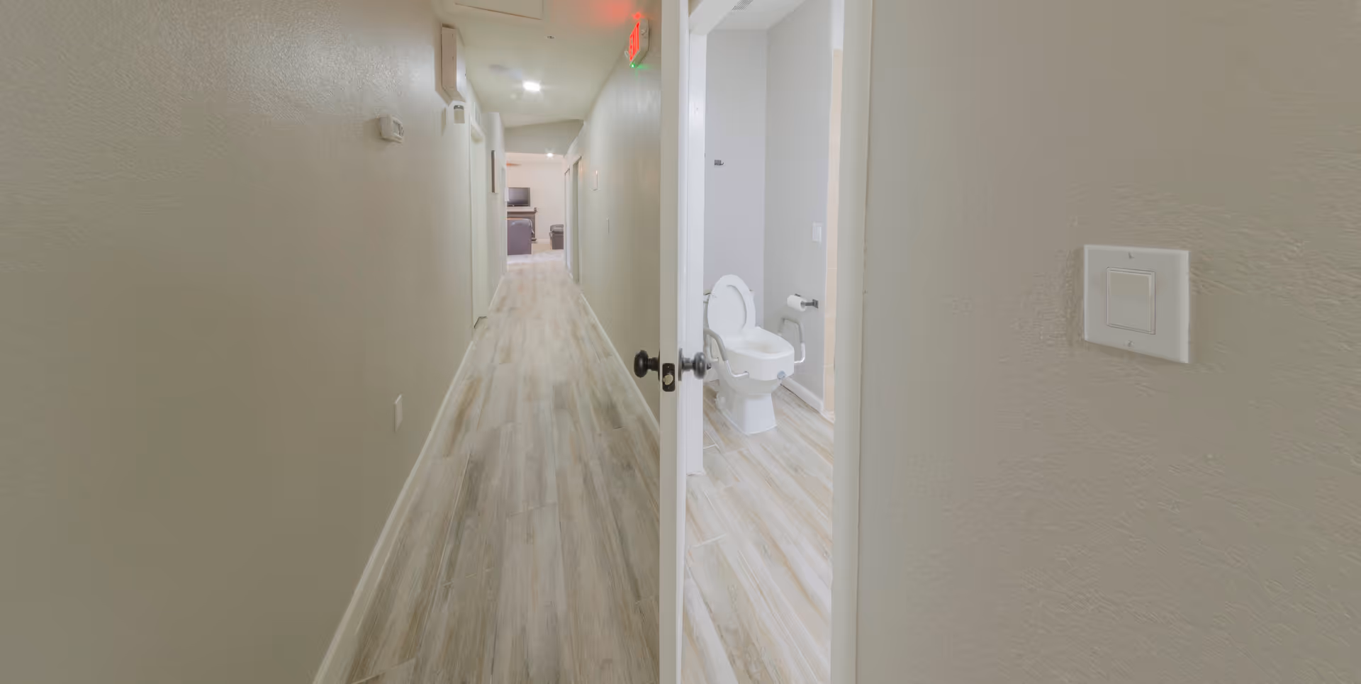 A long hallway with light-colored wood flooring and beige walls. On the right side, there is an open door leading to a bathroom with a toilet equipped with safety rails. At the end of the hallway, a living area with a TV and furniture is visible.