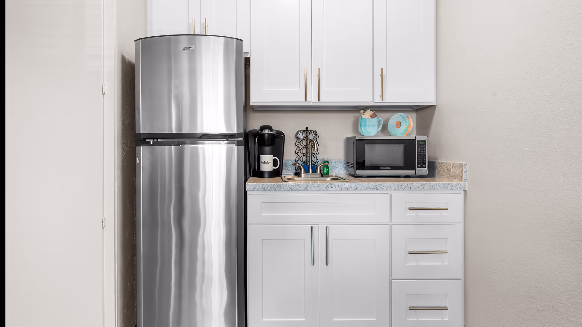 A small kitchen area with a stainless steel refrigerator, white cabinets, a countertop with a coffee maker, a microwave, a small sink, and a few decorative items on the wall.