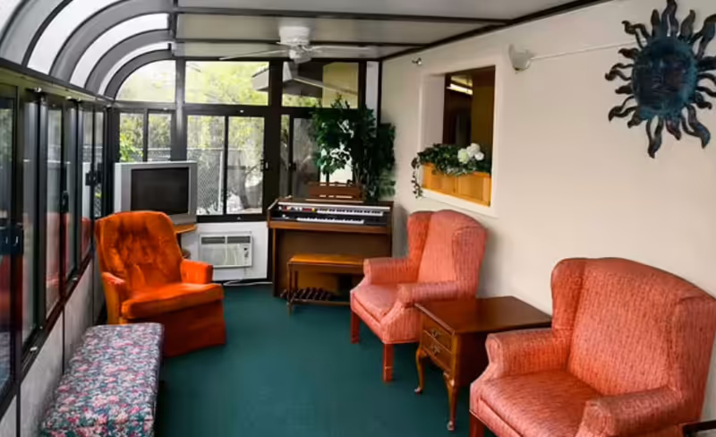 A sunroom with large windows and a curved glass ceiling. The room contains three upholstered armchairs in shades of orange and red, a floral-patterned bench, a wooden side table, a vintage television, an air conditioning unit, a keyboard organ, and a potted plant. A decorative sun wall hanging is mounted on the right wall.