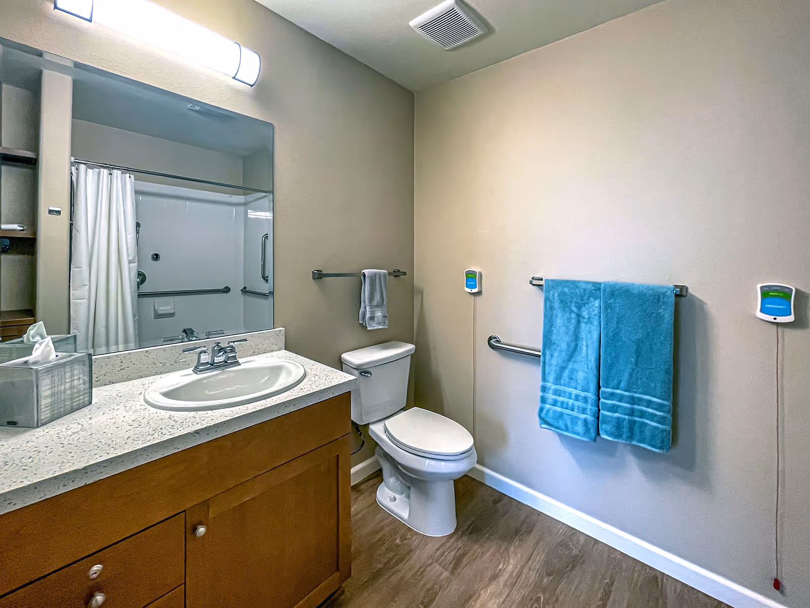 A clean bathroom with a wooden vanity and white sink, a large mirror above the sink, a toilet next to the vanity, and a shower with a white curtain. Two blue towels hang on a towel rack on the wall, and there are two emergency pull cords with green and blue buttons on either side of the towels.