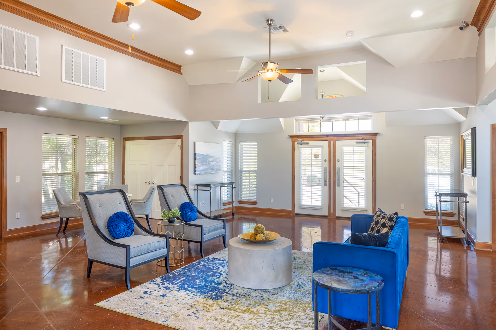 A spacious and well-lit living room area with polished brown floors, a blue velvet sofa with decorative pillows, two gray armchairs with blue round cushions, a round concrete coffee table, and a colorful area rug. The room has large windows with blinds, double glass doors, ceiling fans with lights, and light gray walls with wooden trim.