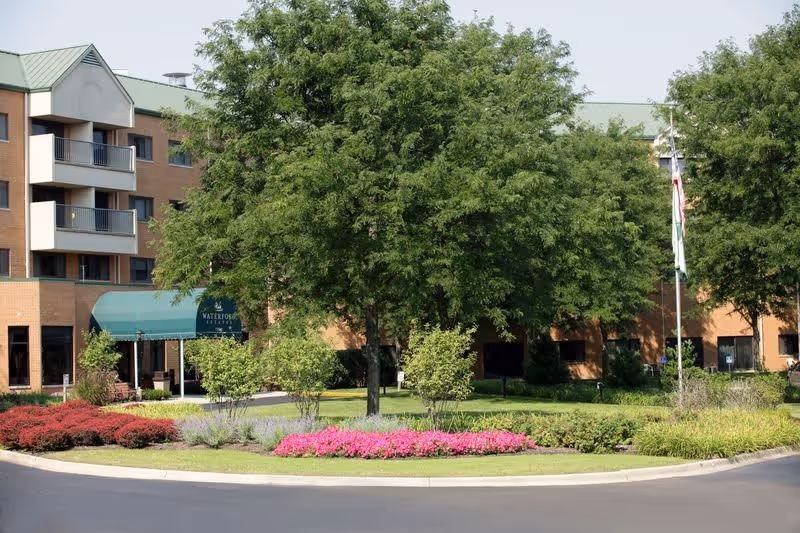 Exterior view of Waterford Estates senior living facility showing a multi-story brick building with balconies, a green awning over the entrance, well-maintained landscaping with trees, bushes, and colorful flowers, and a paved driveway in front.