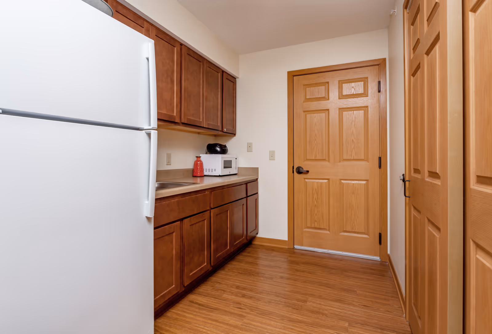 A small kitchen area with wooden cabinets, a white refrigerator, a microwave, a red decorative bottle, and a wooden door. The floor is wood, and the walls are light-colored.