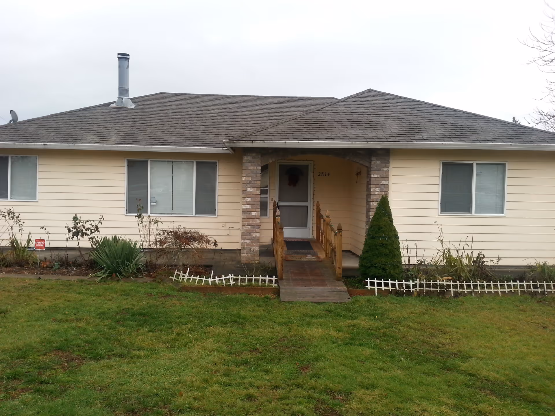 Front exterior view of a single-story beige house with a gray shingled roof, a small wooden ramp leading to the front door, two large windows on either side of the entrance, a small garden with plants and bushes, and a green lawn in the foreground under an overcast sky.