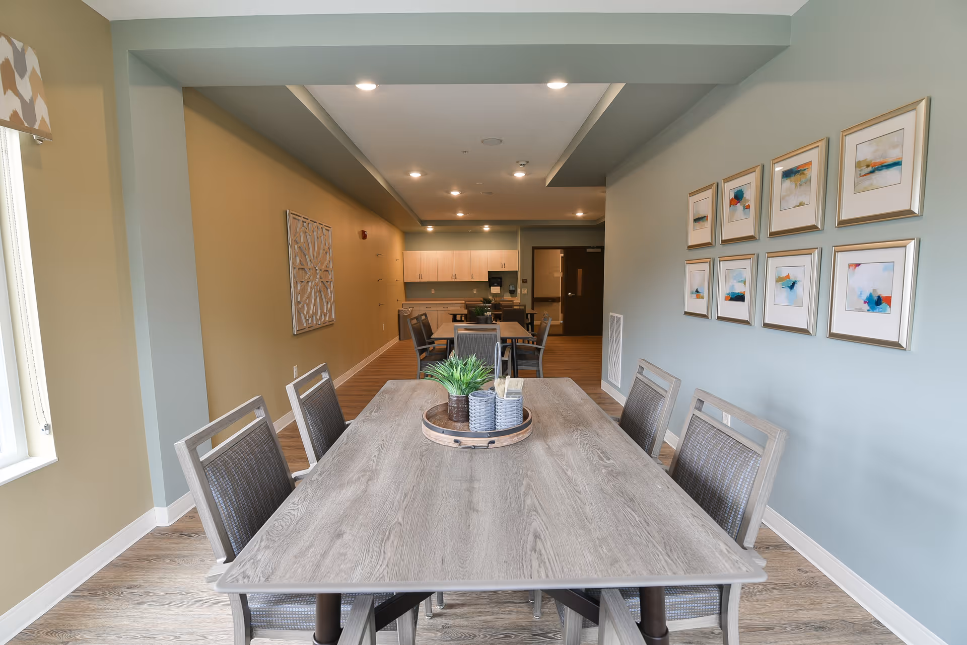 Communal dining room with a long wooden table and chairs, framed artwork on the wall, and a kitchenette area visible in the background.