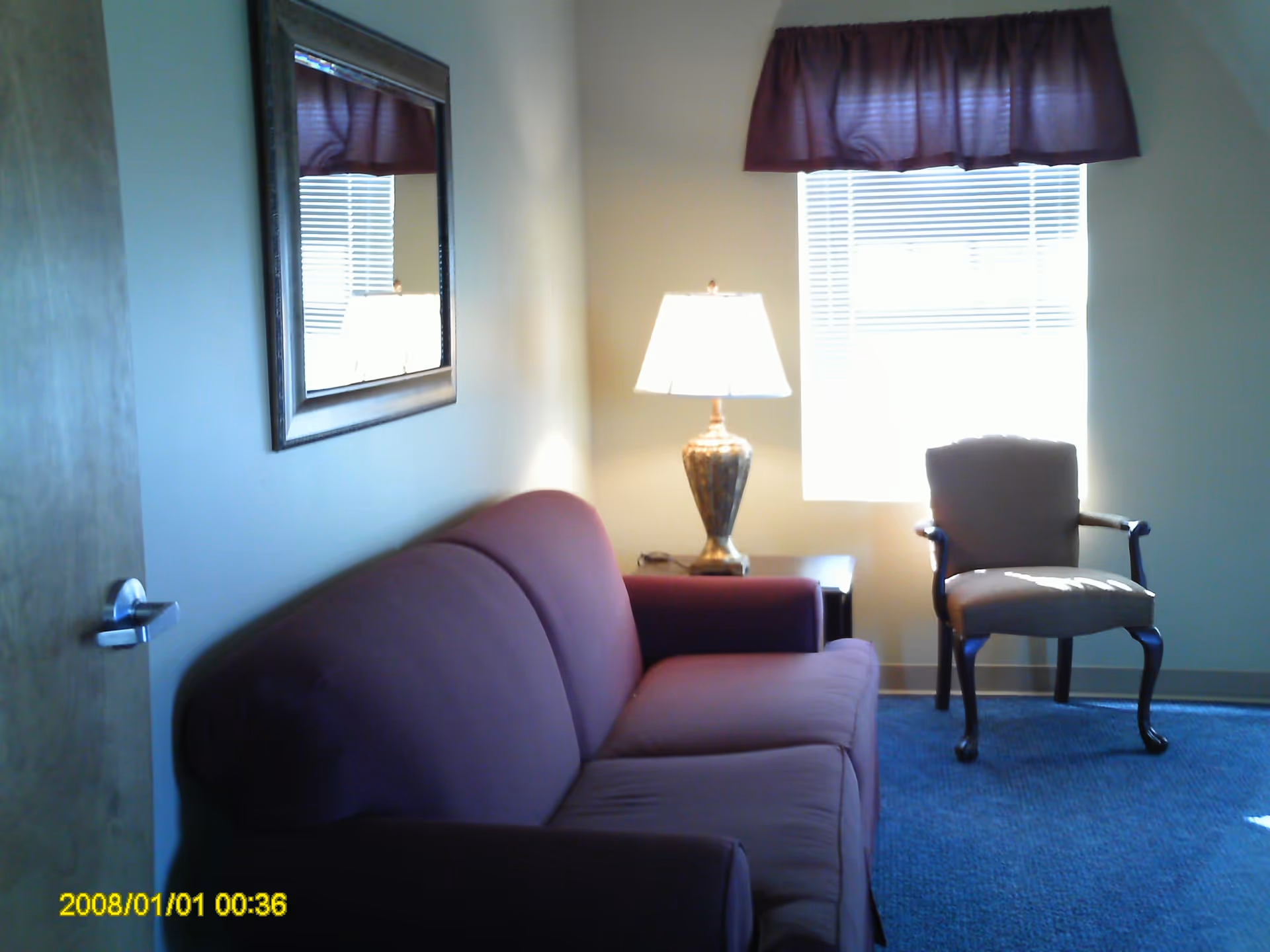A small living room with a maroon sofa on the left, a framed mirror above it, a wooden side table with a decorative lamp, and a single upholstered armchair near a window with blinds and a maroon valance.