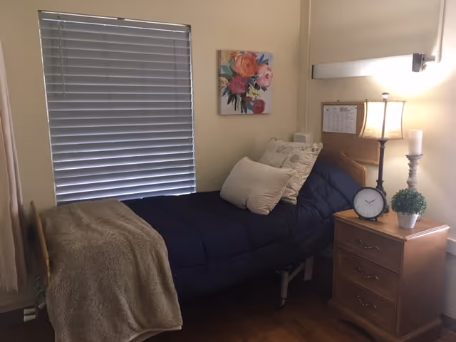 A small bedroom in a nursing home with a single bed covered in a dark blue comforter and beige pillows. A beige blanket is draped over the foot of the bed. Next to the bed is a wooden nightstand with three drawers, holding a lamp, a clock, a small plant, and a candle. A floral painting hangs on the wall above the bed, and a window with closed blinds is on the left side.