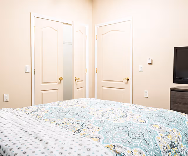 A bedroom with a bed covered in a patterned quilt featuring blue, yellow, and white colors. The room has beige walls, two closed doors with gold handles, and a flat-screen TV on a dark wooden dresser.