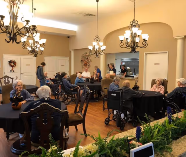 A group of elderly people, some in wheelchairs, sitting around several round tables covered with black tablecloths in a warmly lit dining room. There are chandeliers hanging from the ceiling, and a few staff members are standing near a serving area in the background. The room has beige walls and wooden flooring, with decorative plants in the foreground.