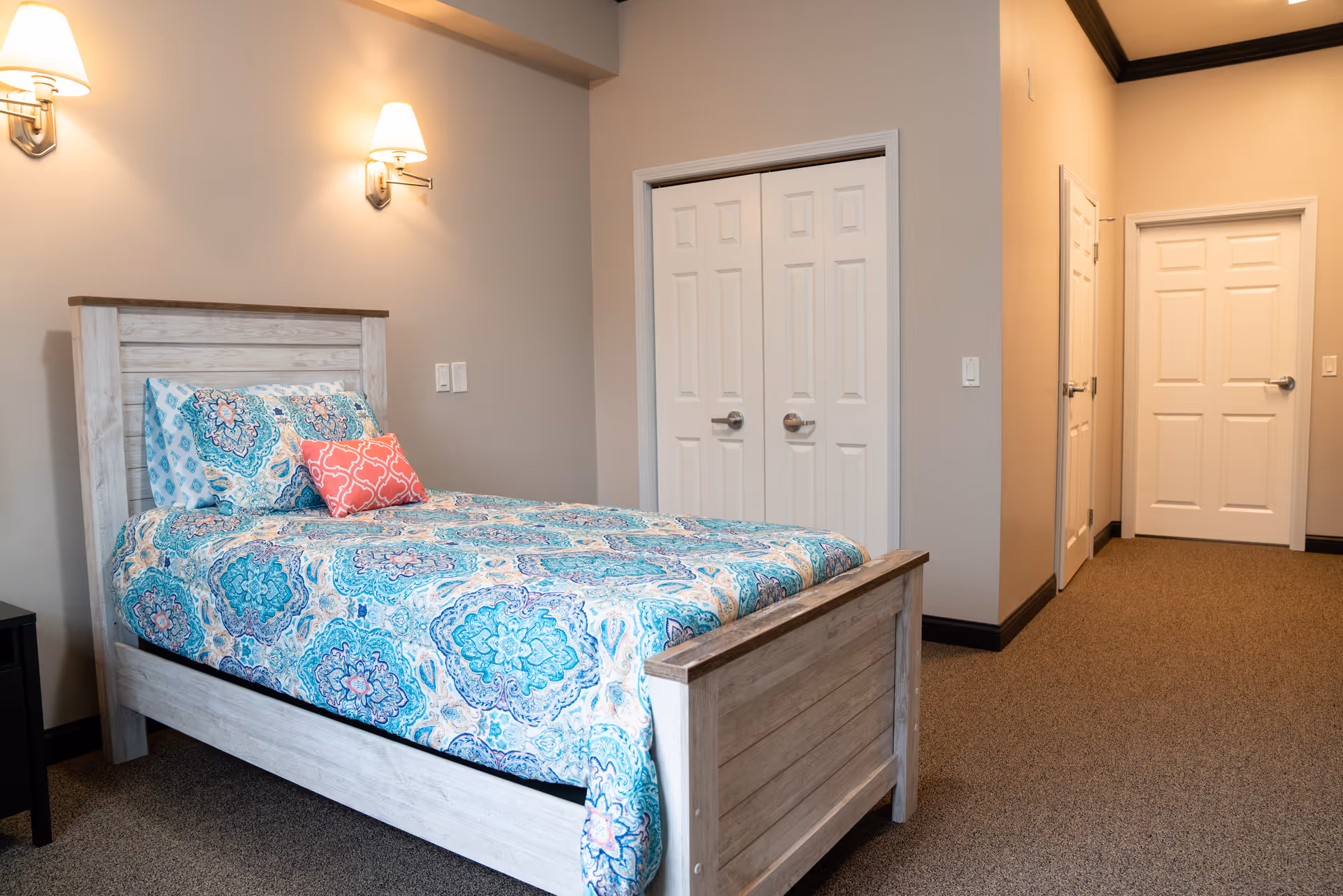A neatly made single bed with patterned blue bedding in a furnished bedroom with wall sconces and closet doors.