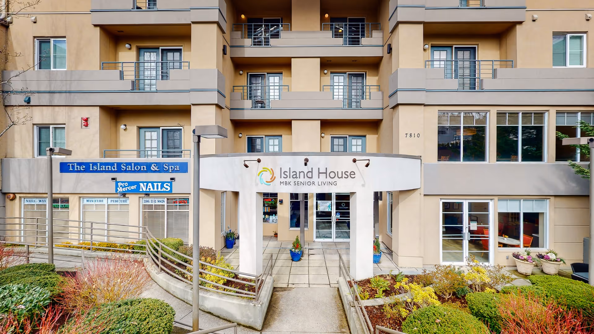 Front exterior view of Island House MBK Senior Living building with entrance canopy, balconies, and landscaped garden beds. Adjacent to the entrance is The Island Salon & Spa with a blue sign and a Pro Mercer Nails sign.