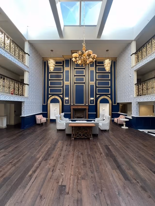 A spacious and elegant living room area with dark wood flooring, a high ceiling with skylights, and ornate gold and navy blue wall paneling. The room features a central seating arrangement with white upholstered chairs around a wooden coffee table, a fireplace, two sculptures in alcoves, and a large chandelier hanging from the ceiling. There are also balconies with decorative railings on both sides of the room.