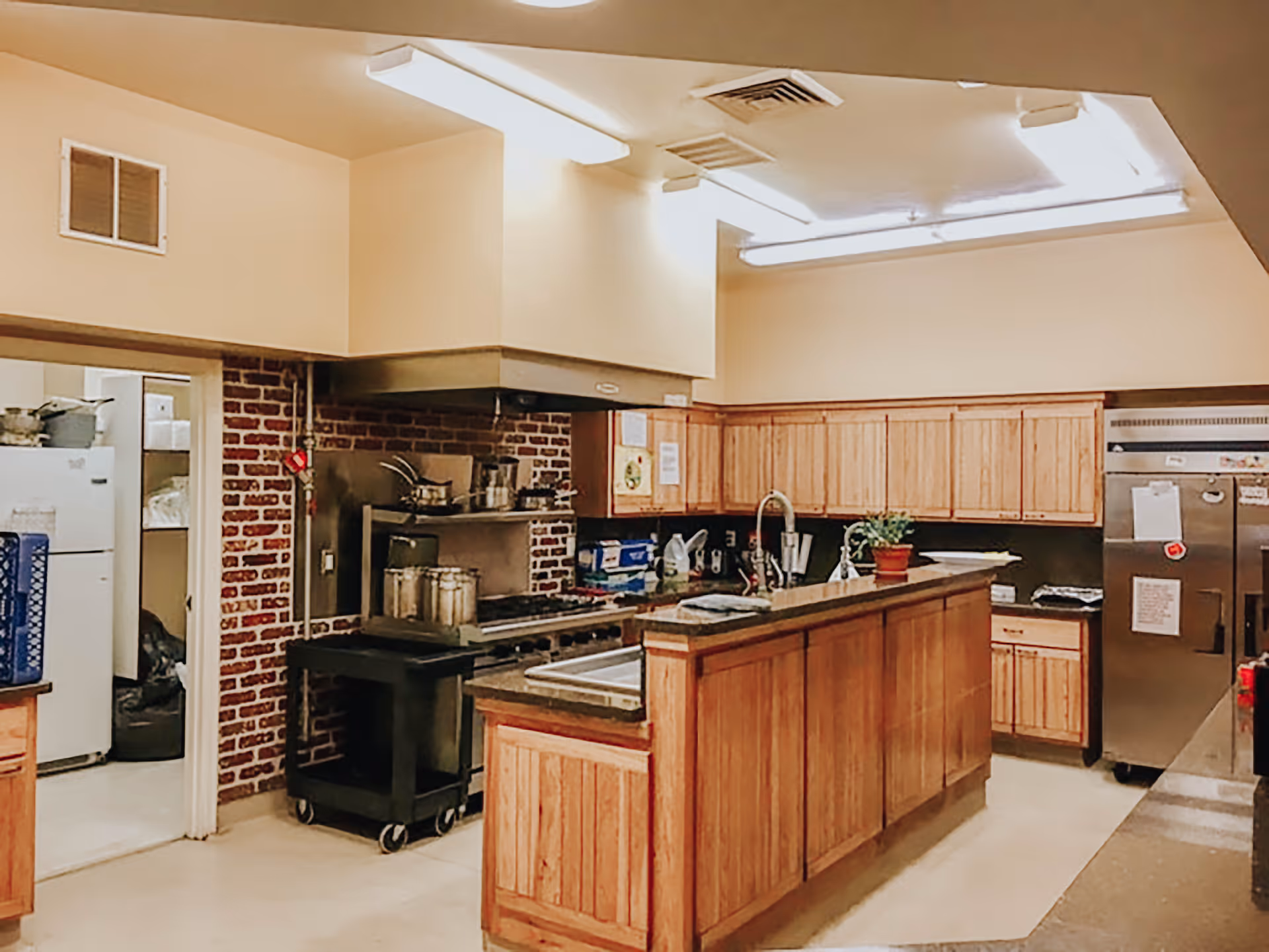 Interior view of a kitchen with wooden cabinets, a large stainless steel refrigerator, a stove with pots, a sink on a central island, and bright overhead fluorescent lighting.