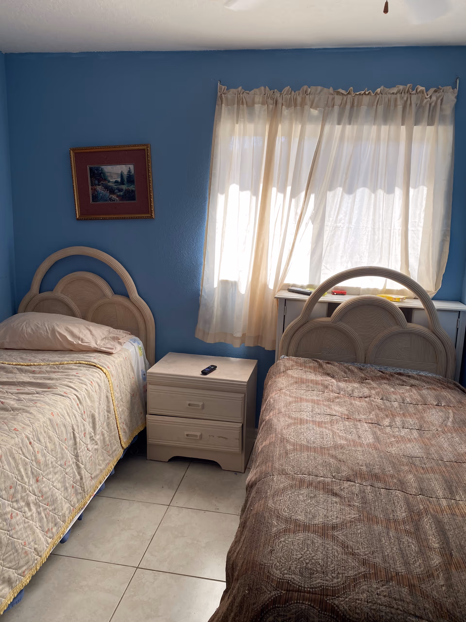 A small bedroom with two single beds separated by a light-colored nightstand with two drawers. Each bed has a different patterned bedspread, one beige and the other brown. A window with sheer white curtains lets in natural light, and a framed painting hangs on the blue wall above one of the beds.