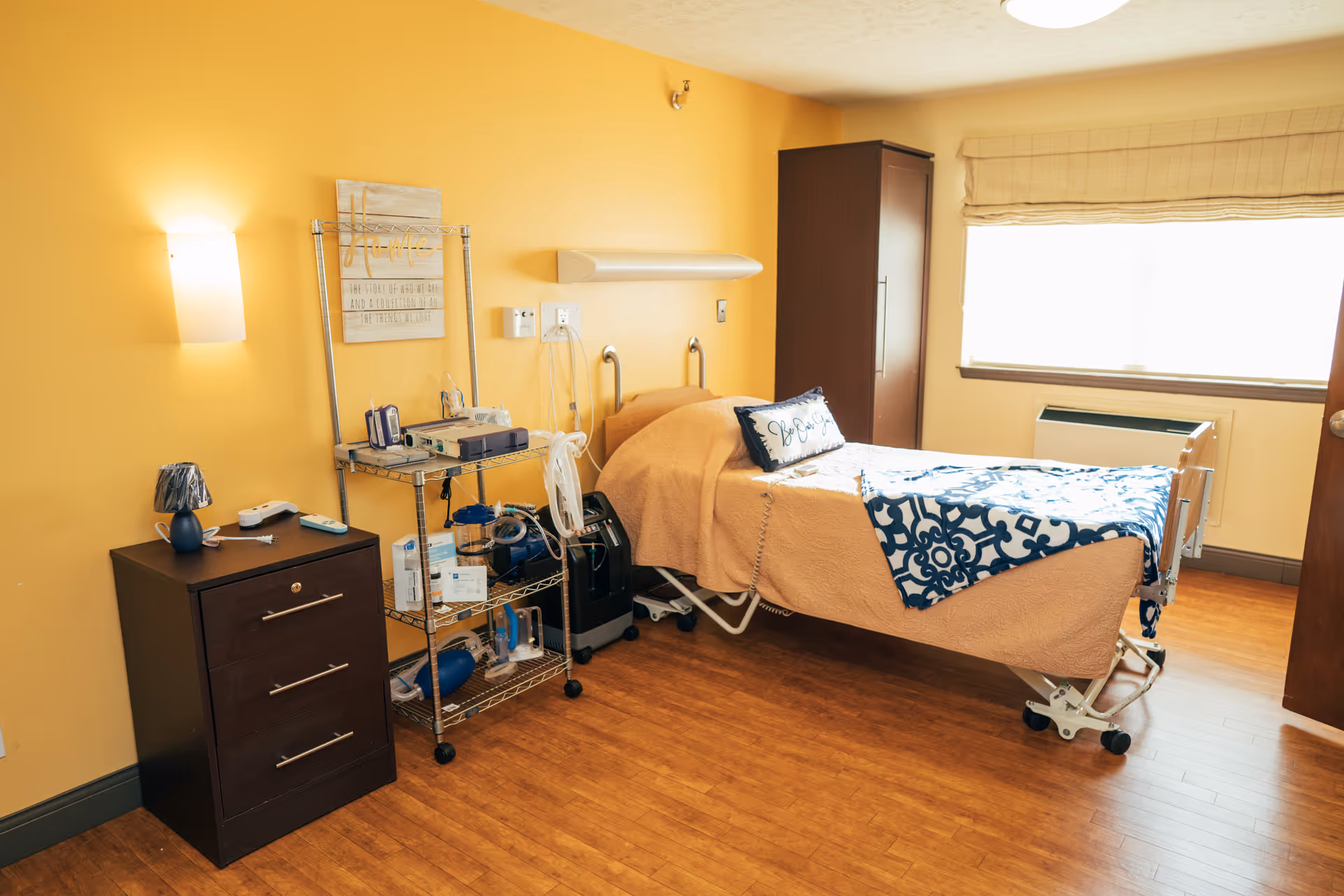 A well-lit rehabilitation room with a single hospital-style bed covered in a beige quilt and a blue and white patterned blanket. The bed has a decorative pillow with the words 'Be Joyful'. Next to the bed is a metal rack holding medical equipment and supplies. A dark wooden nightstand with three drawers and a small lamp is positioned against a yellow wall. A window with a beige Roman shade allows natural light into the room, and a tall dark wooden cabinet stands near the window. The floor is wooden, and the room has a warm, inviting atmosphere.