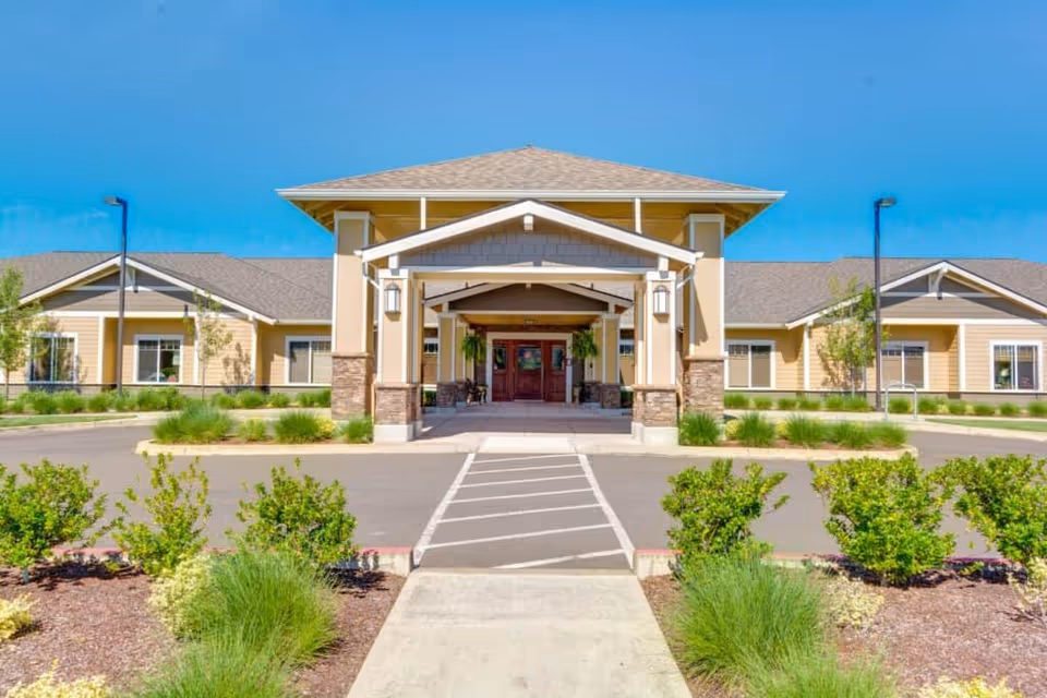 Front exterior view of Arbor Oaks Terrace Memory Care Residence building with a covered entrance, beige siding, stone accents, and landscaped greenery under a clear blue sky.