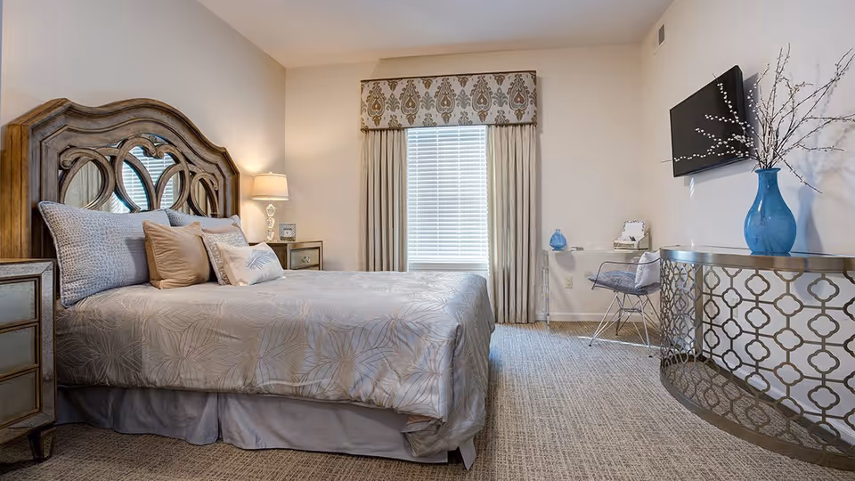 A well-decorated bedroom with a large wooden headboard bed covered in light-colored bedding and multiple pillows. To the left of the bed is a nightstand with a lamp and a small picture frame. A window with closed blinds and patterned curtains is centered on the back wall. On the right side of the room, there is a decorative console table with a blue vase holding branches, a clear acrylic chair with a cushion, and a wall-mounted flat-screen TV above the table. The room has neutral-colored walls and carpeted flooring.