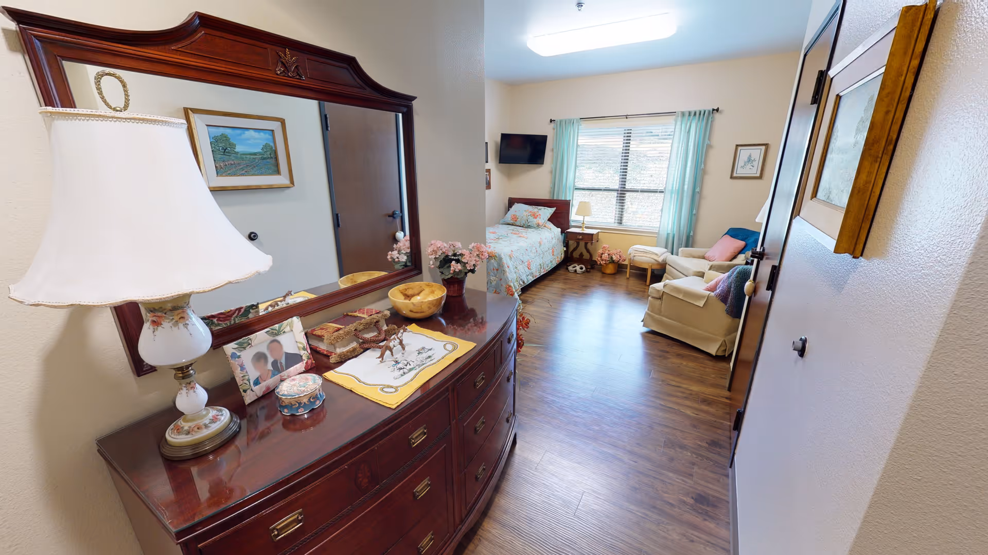 A cozy senior living bedroom with a wooden dresser topped with a decorative lamp, framed photos, and small ornaments. The room features a single bed with floral bedding, a small side table with a lamp, a comfortable armchair with a pillow and blanket, and a window with light blue curtains letting in natural light. The floor is wooden, and the walls are decorated with framed pictures.