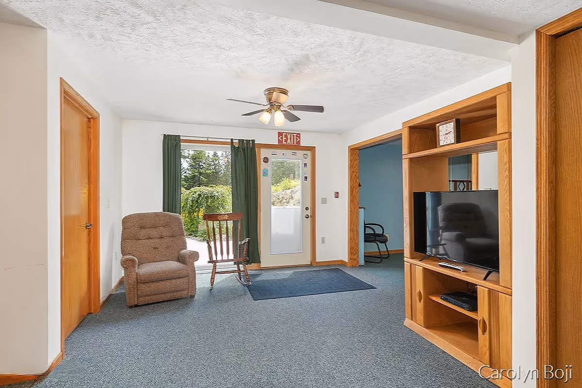 A cozy living room area with a brown upholstered armchair and a wooden rocking chair near a glass door with green curtains. The door has an exit sign above it and leads outside to greenery. To the right, there is a wooden entertainment center with a flat-screen TV and a clock on the top shelf. The room has blue carpet and white walls with wooden trim, and a ceiling fan with lights is mounted on the ceiling.
