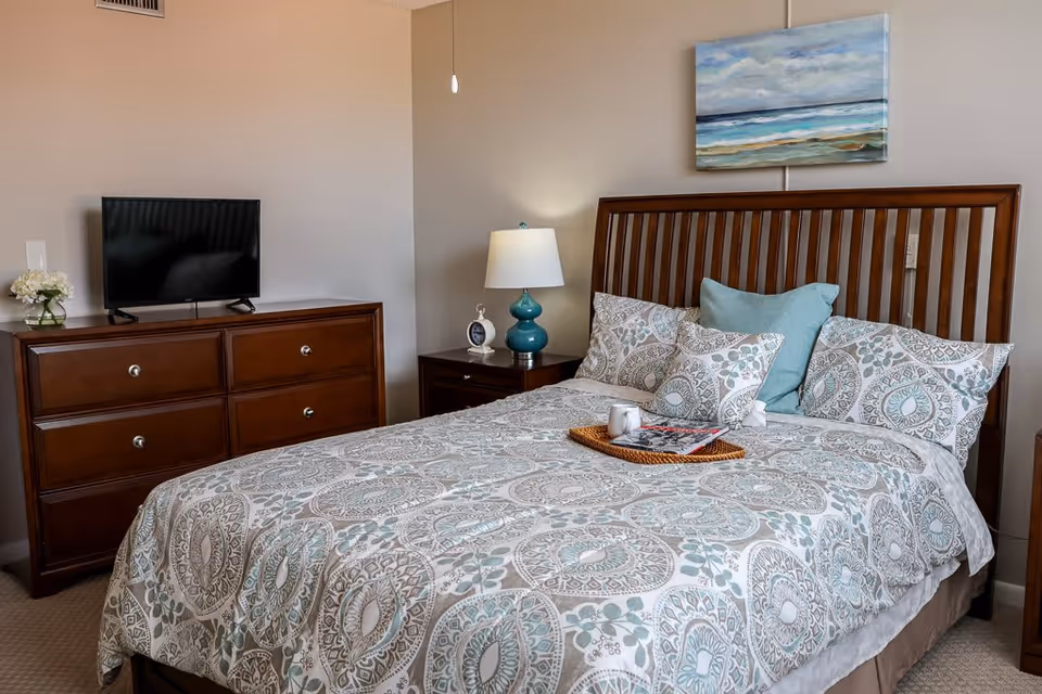 A neatly made bed with patterned bedding and multiple pillows in a bedroom. Next to the bed is a wooden nightstand with a blue lamp and a small clock. Across from the bed is a wooden dresser with a flat-screen TV and a small vase of flowers. A painting of a seascape hangs on the wall above the bed.