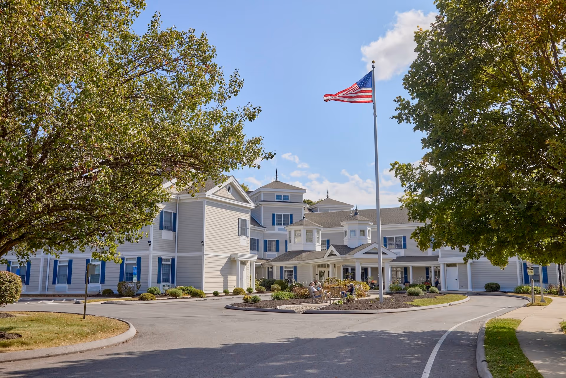 Exterior view of Monarch Southbury senior living facility on a sunny day, featuring a large building with white siding and blue shutters, an American flag on a tall flagpole in front, surrounded by trees and a circular driveway with some people sitting near the entrance.