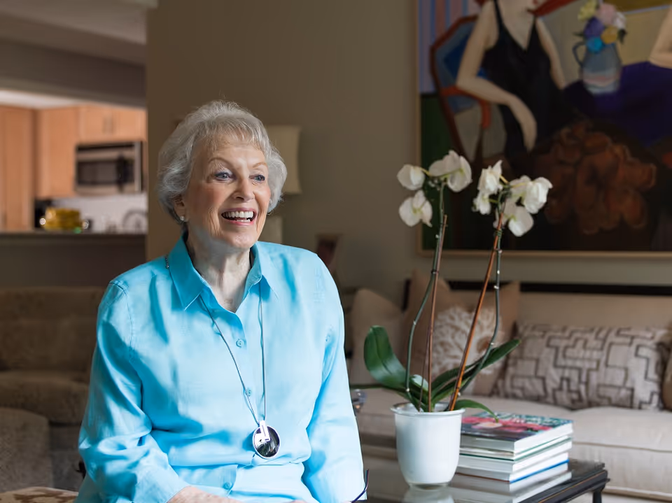 An elderly woman with short gray hair wearing a light blue shirt is sitting and smiling in a living room. Behind her is a beige couch with patterned pillows, a coffee table with a white orchid plant and stacked books, and a colorful painting on the wall. The background also shows a kitchen area with wooden cabinets and a microwave.
