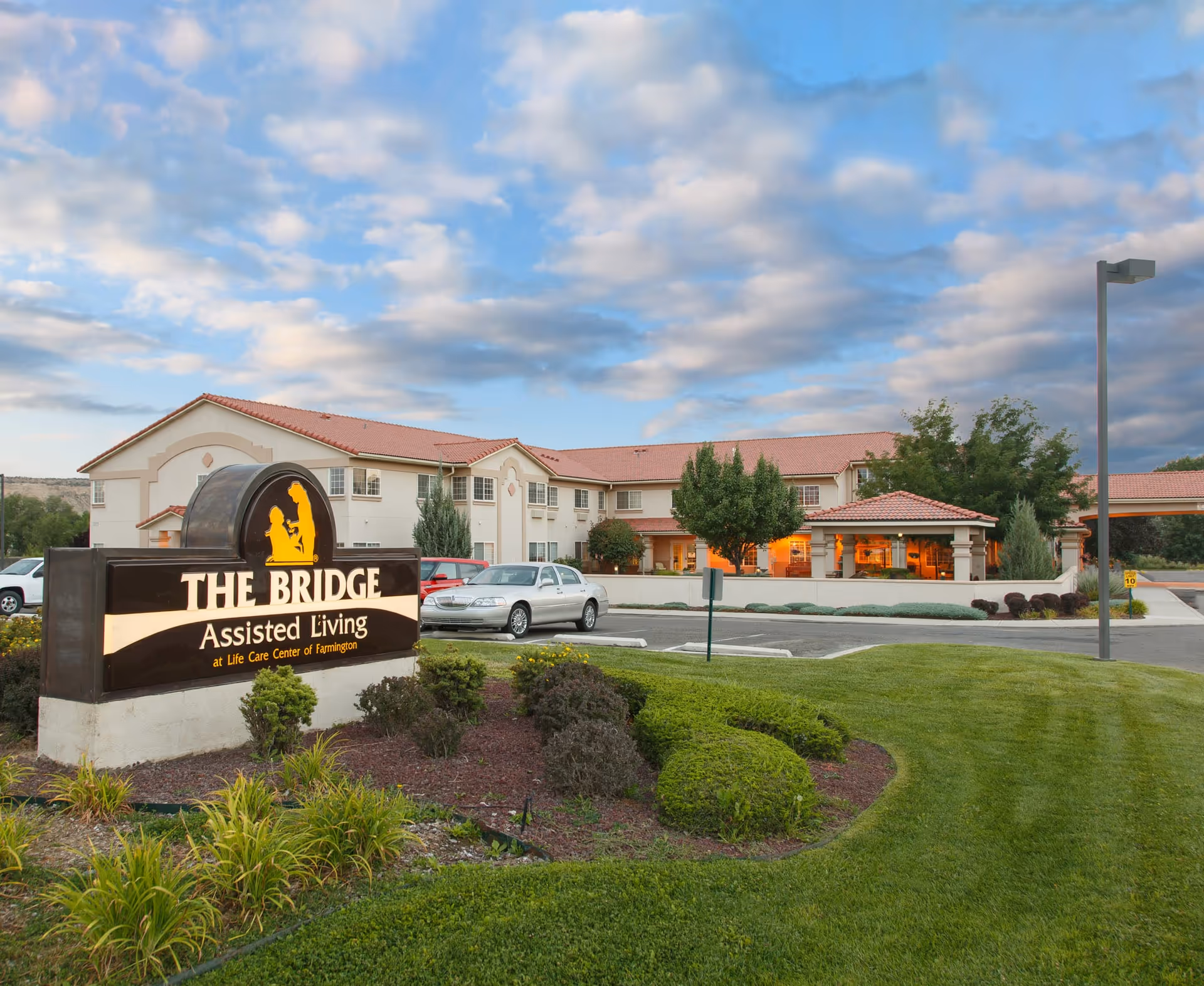 Exterior view of The Bridge at Farmington Assisted Living facility with a large sign in the foreground, landscaped greenery, parked cars, and a two-story building under a partly cloudy sky.
