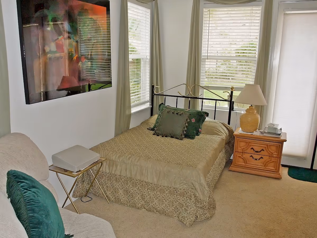 A cozy bedroom with a bed covered in a beige patterned bedspread and green decorative pillows. The room has two large windows with white blinds and light green curtains. There is a wooden nightstand with a beige lamp and a tissue box next to the bed. A beige armchair with a green pillow and a small side table with a white electronic device are also visible. A colorful abstract painting hangs on the wall above the side table.