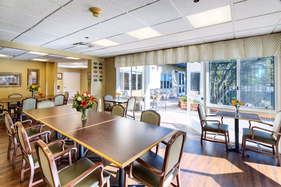 Bright memory care dining room with several tables and chairs, vases of flowers, and large windows/doors opening to an outdoor patio.