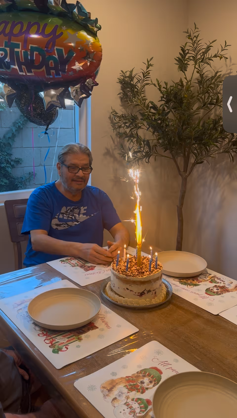 An elderly man wearing glasses and a blue Nike t-shirt sits at a dining table with a birthday cake in front of him. The cake has lit candles and a sparkler. There are empty plates and festive placemats with holiday designs on the table. A large 'Happy Birthday' balloon is visible near a window, and a tall indoor plant is in the background.